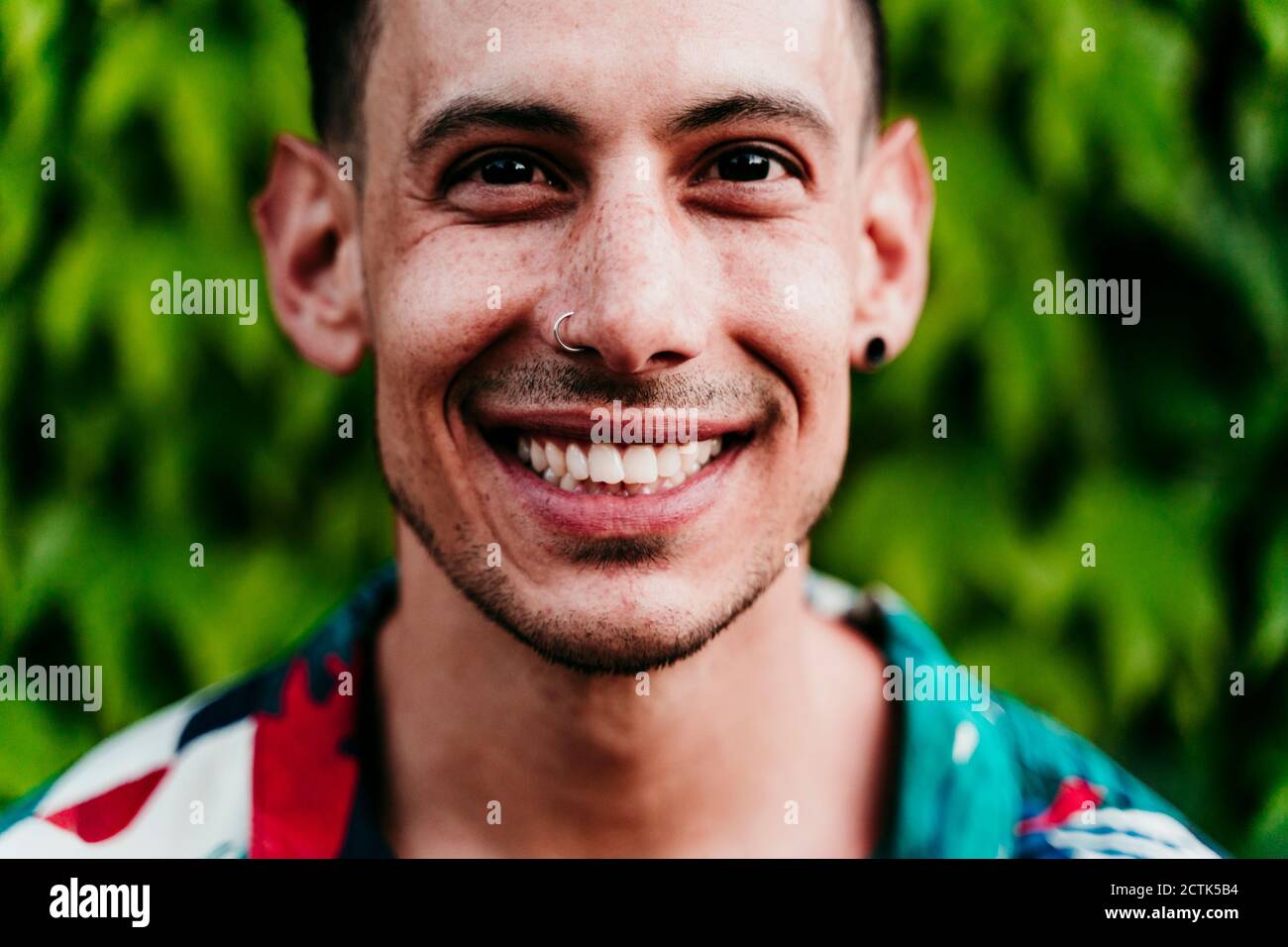 Man wearing nose ring smiling while standing against ivy hedge Stock Photo