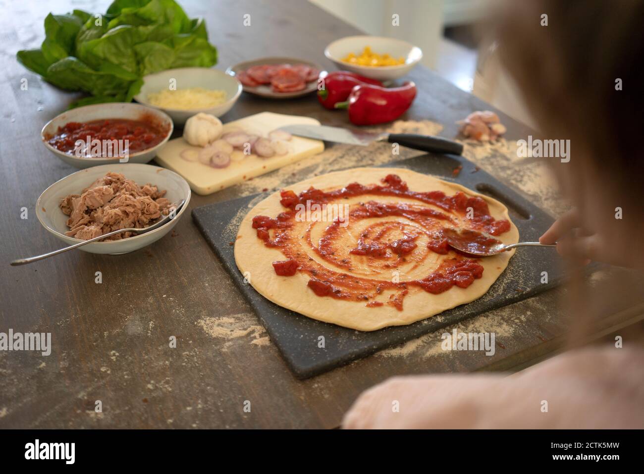 Girl spreading sauce over dough while preparing pizza on kitchen island Stock Photo