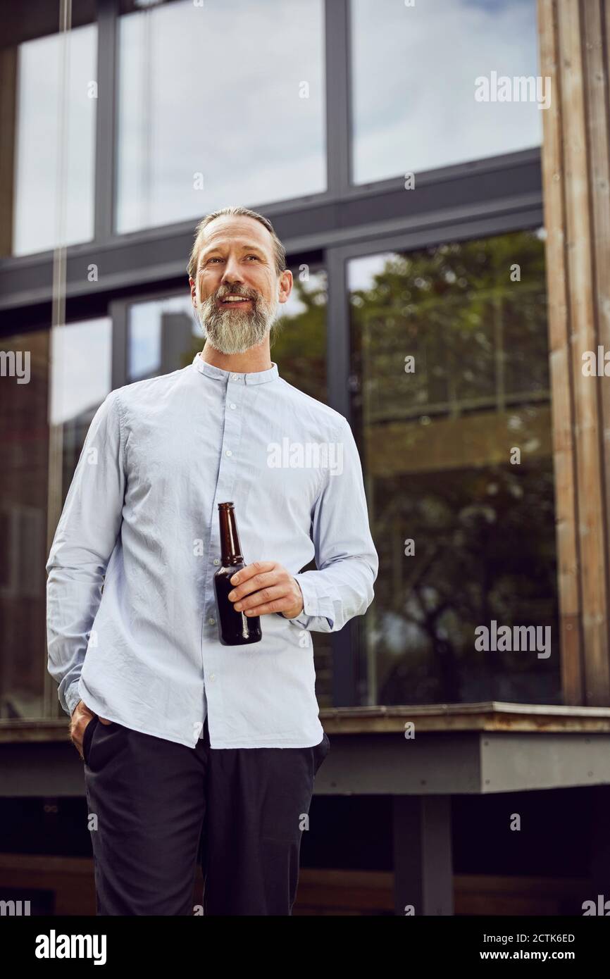 Bearded mature man holding beer bottle while standing against tiny house Stock Photo