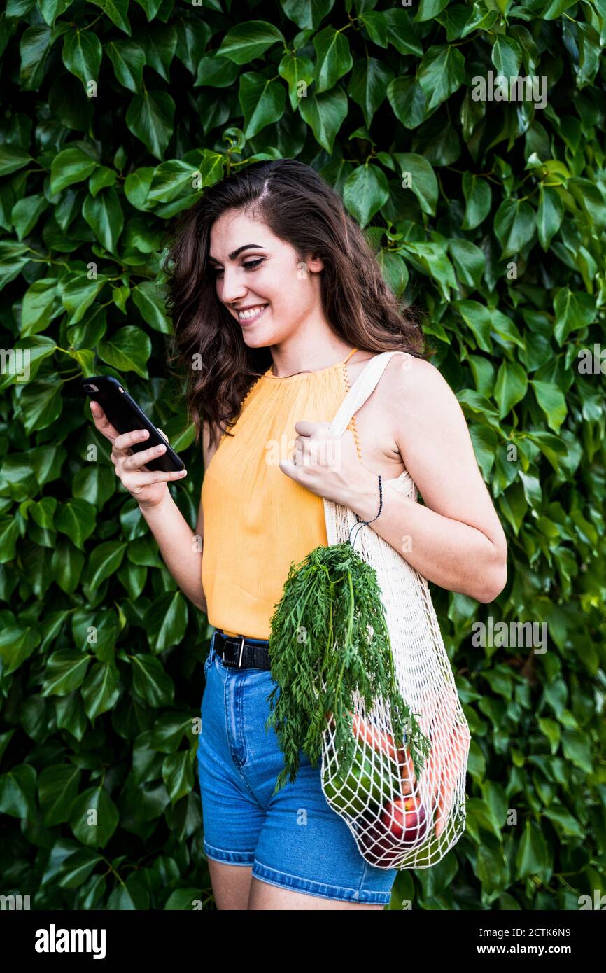 Happy young woman using smart phone while standing with vegetables in mesh bag against green plants Stock Photo