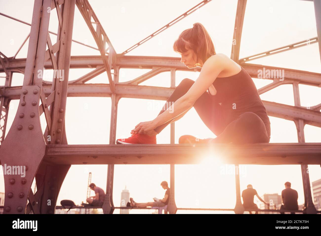 Woman tying shoelace while sitting on metal bridge at sunset Stock Photo
