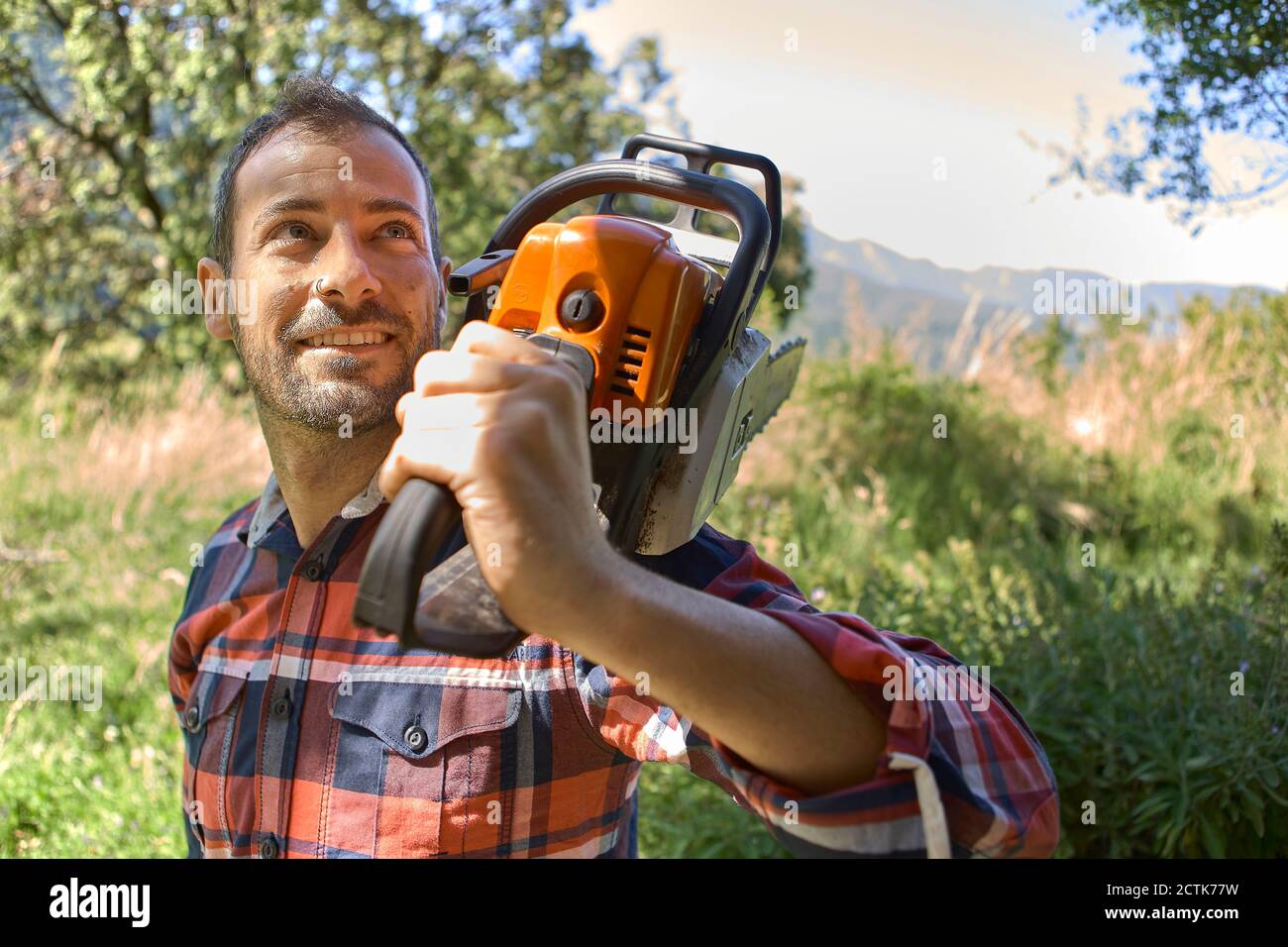 Smiling male lumberjack carrying electric saw on shoulder in forest Stock Photo