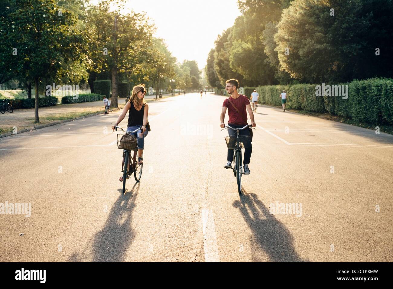 Happy couple cycling on road amidst trees in park during sunset Stock Photo