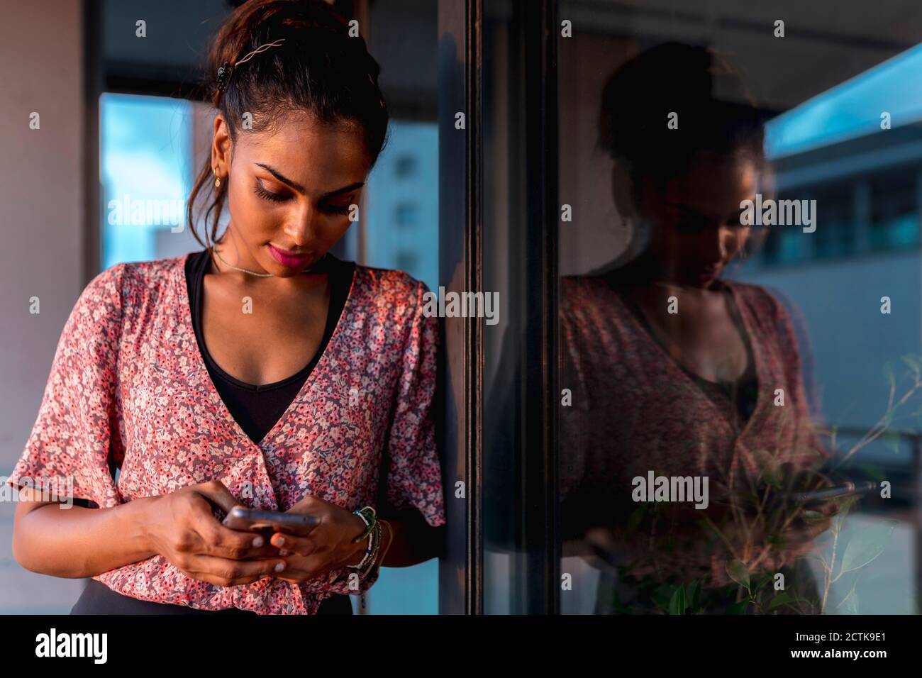 Beautiful woman using phone while leaning by reflection on window in balcony Stock Photo