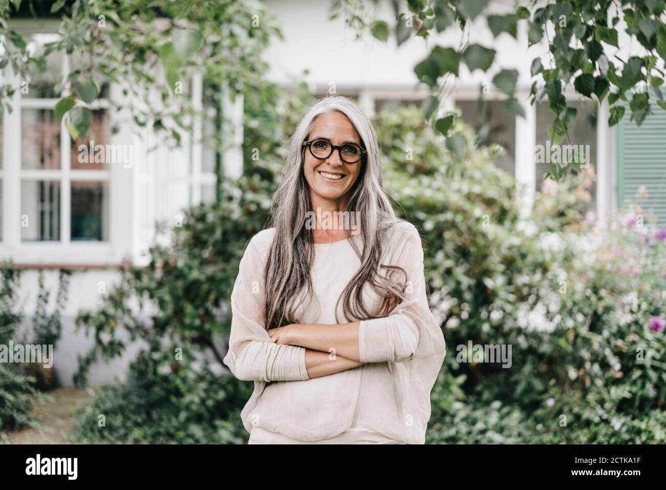 Portrait of smiling woman with long grey hair wearing spectacles standing in the garden Stock Photo