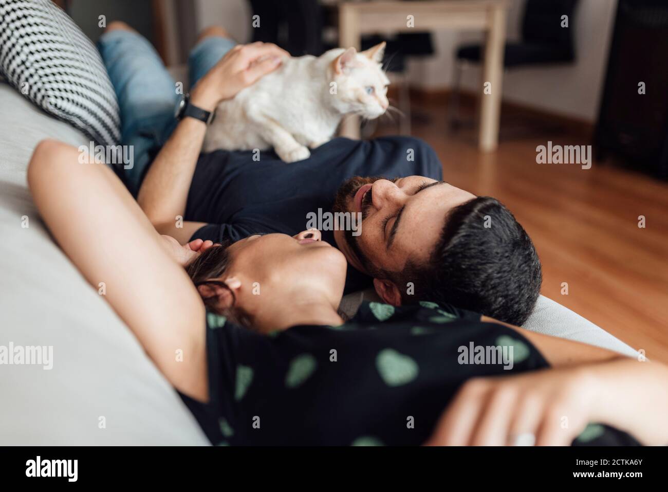 Young woman and mid adult man lying down on sofa shares tenderness with cat at home Stock Photo