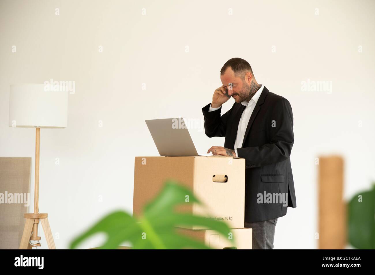 Male entrepreneur working over laptop on stacked boxes against wall in new home Stock Photo