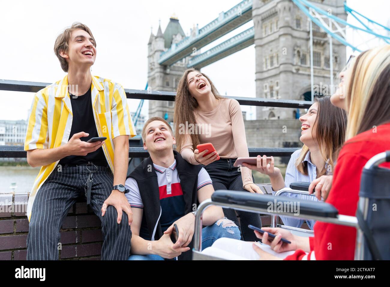 Cheerful male and female friends with mobile phones sitting against Tower Bridge, London, UK Stock Photo