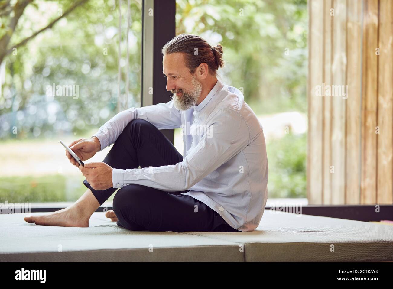 Smiling bearded mature man using digital tablet while sitting on bed by window at home Stock Photo