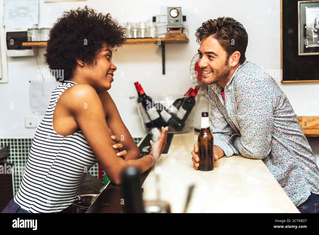 Smiling man holding beer bottle flirting with female bartender while standing at bar counter