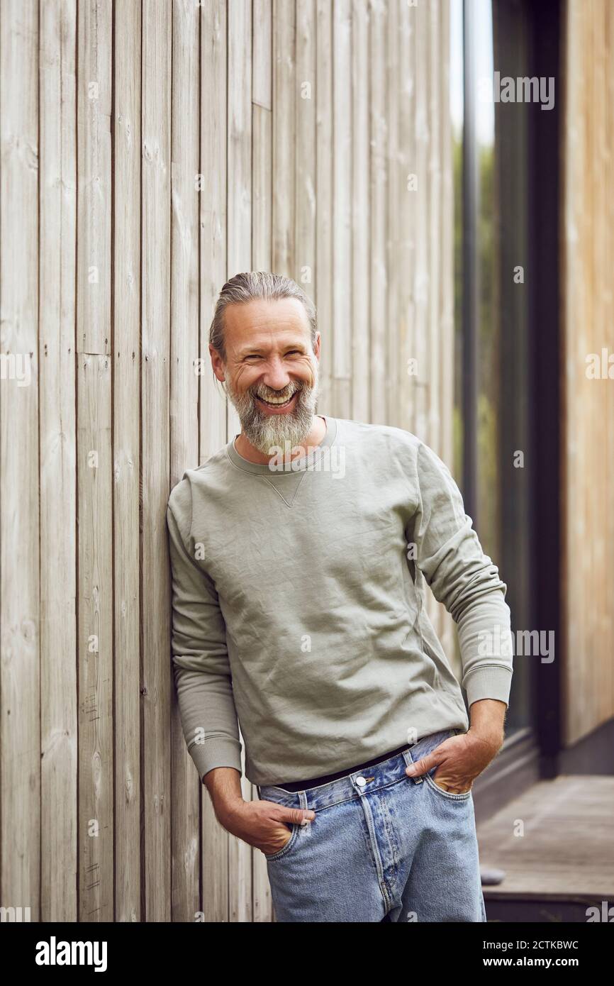 Happy bearded mature man with hands in pockets standing by wall at yard Stock Photo