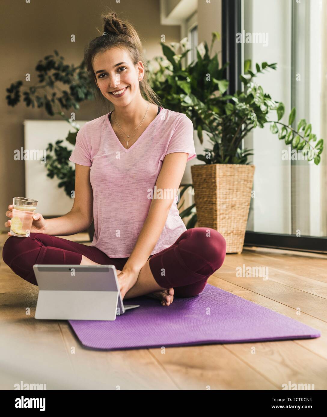 Smiling young woman with water and digital tablet sitting on exercise mat at home Stock Photo