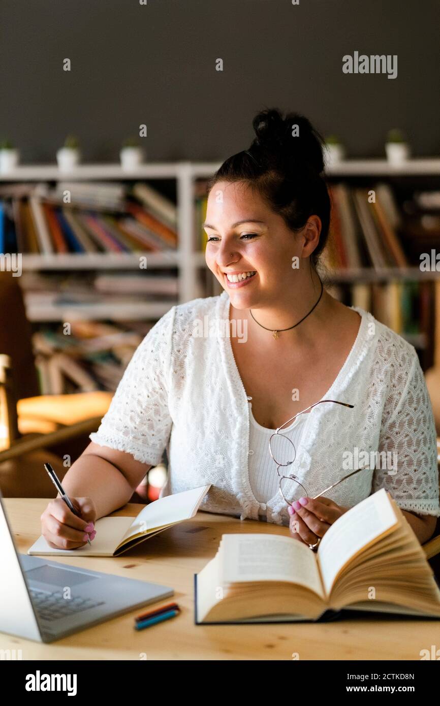 Smiling young woman writing in book while studying over laptop on table at coffee shop Stock Photo
