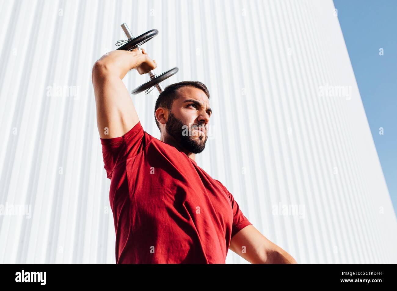 Young bearded man lifting dumbbell up while standing against wall Stock Photo