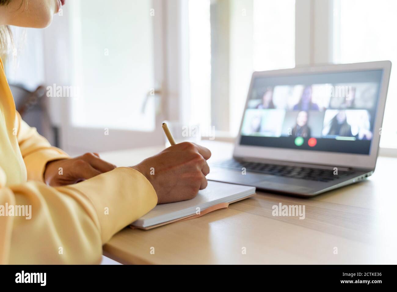 Cropped hands of female entrepreneur writing on diary while talking with colleagues through video conference at home Stock Photo