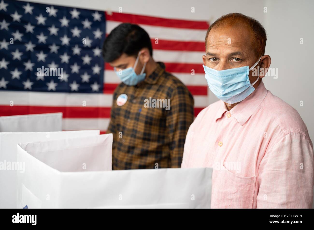 People with mask busy at polling booth for voting with US flag as background - concept of in person voting with covid-19 safety measures at us Stock Photo