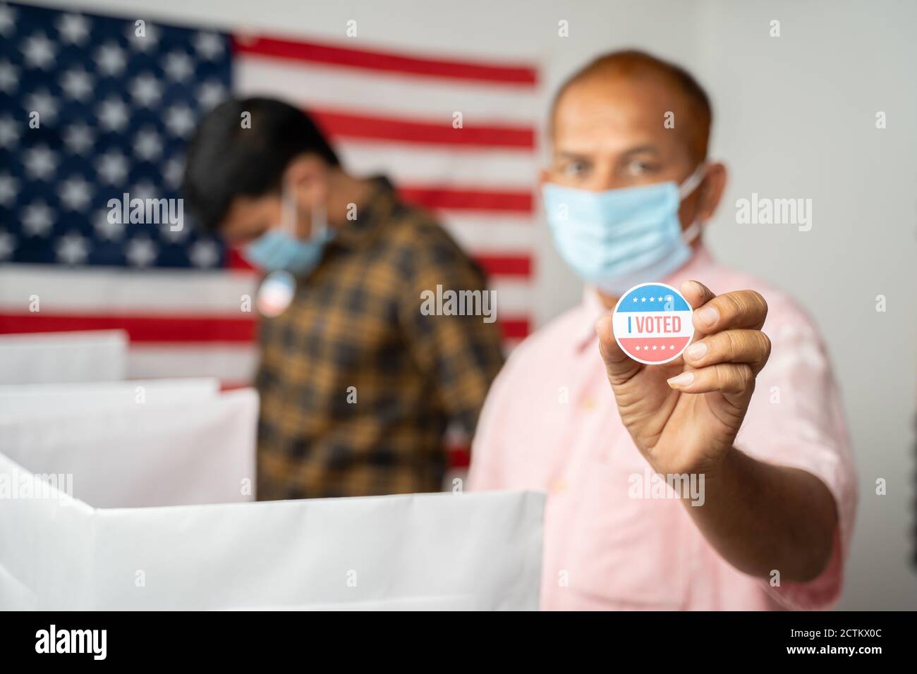 Man in medical mask showing I voted Sticker at polling booth with US flag as background - concept in person voting at US election. Stock Photo