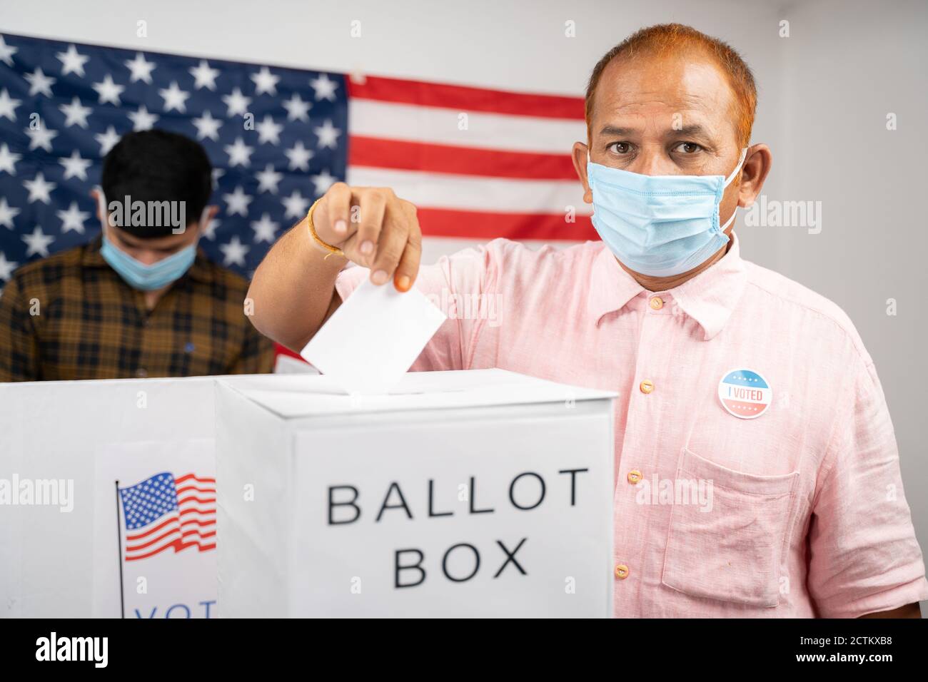 Man in medical mask placing ballot paper inside the ballot box while looking at camera - Concept of in person voting and people busy at polling booth Stock Photo