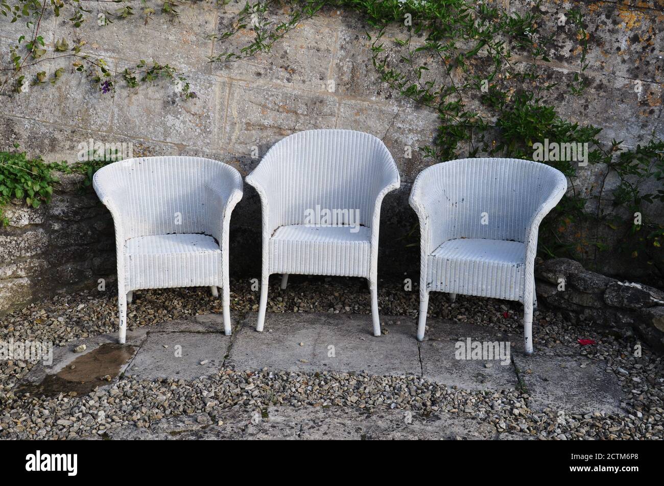 A row of three white wicker Lloyd Loom chairs in a sunken patio of stone chippings and a retaining stone wall in an English country garden. Stock Photo