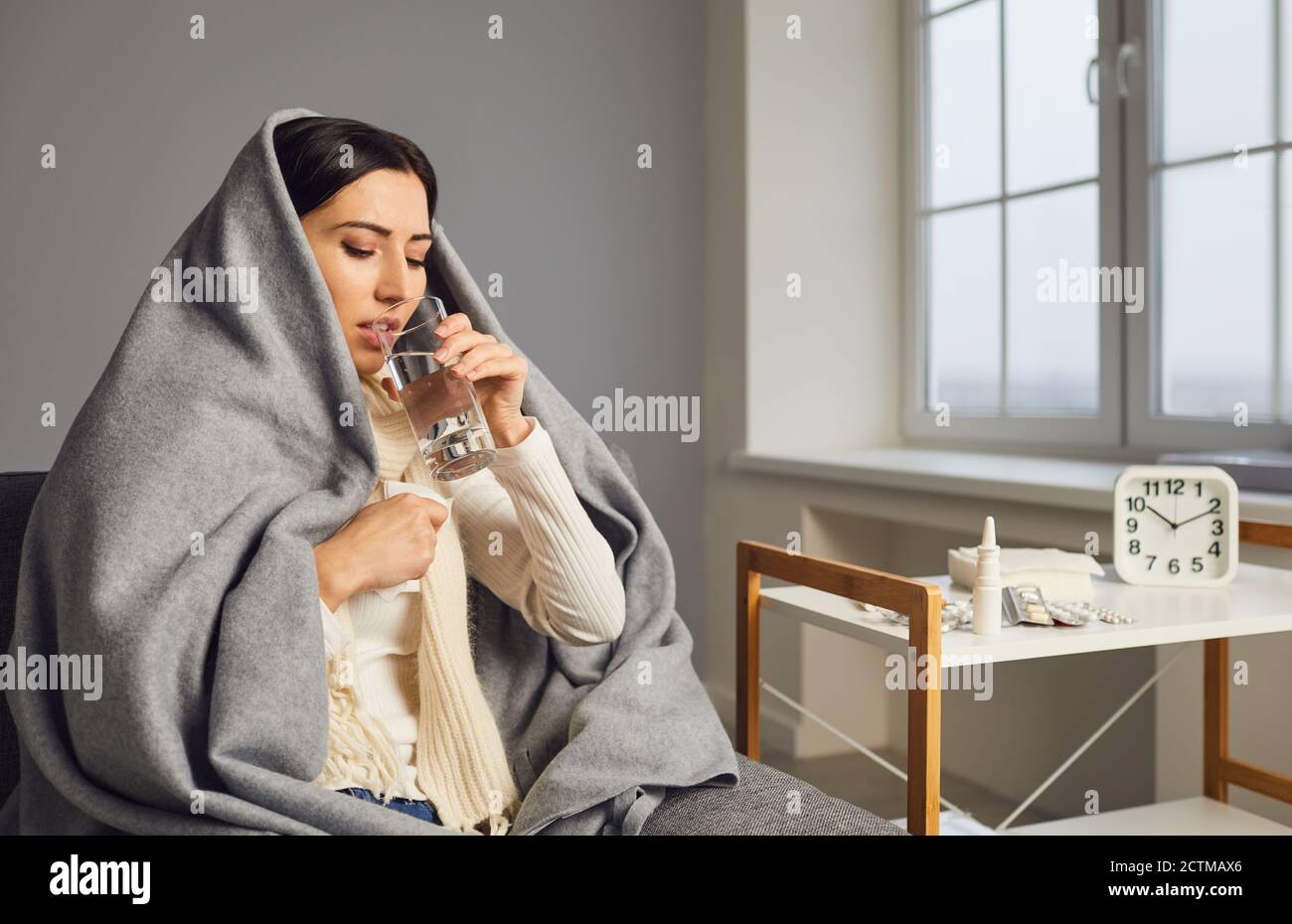 Side view of young woman in blanket caught cold and taking flu medicine, drinking water. Stock Photo