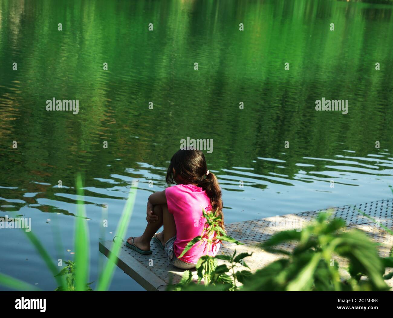 Pensive little girl seen from behind, sitting on a pontoon on the riverside, contemplating nature Stock Photo