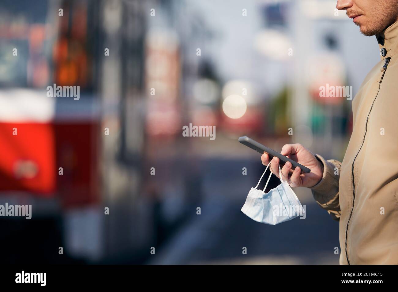 Young man with face mask in hand using smart phone during waiting for tram. Themes modern technology in new normal, coronavirus and personal protectio Stock Photo