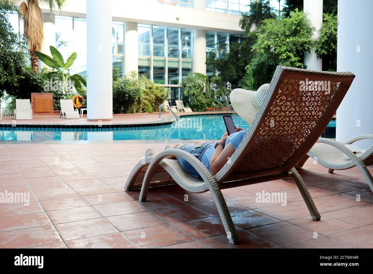Woman in hat lying on a lounge chair with smartphone near the indoor swimming pool. Relax and vacation on resort at autumn or winter Stock Photo