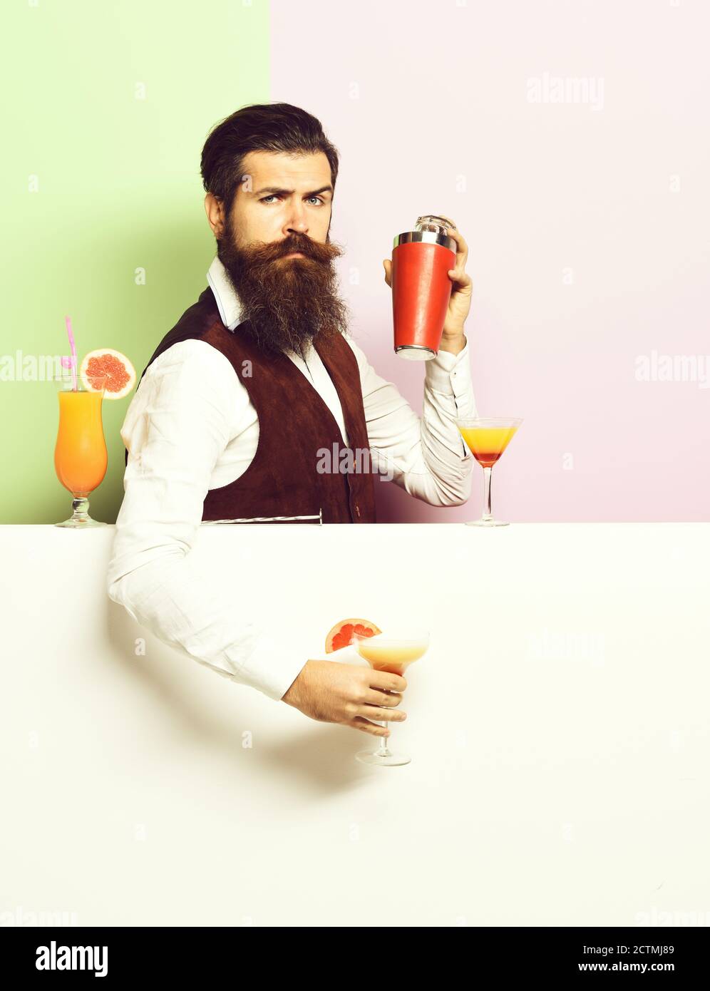 handsome bearded barman with long beard and mustache has stylish hair on serious face holding shaker and made alcoholic cocktail in vintage suede leather waistcoat on purple green studio background Stock Photo