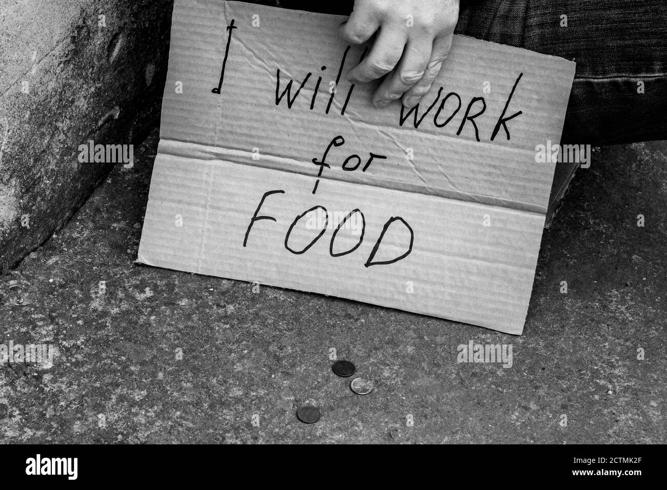 unemployed man sitting on the ground holding a cardboard sign saying i will work for food alms coins on earth black and white image Stock Photo