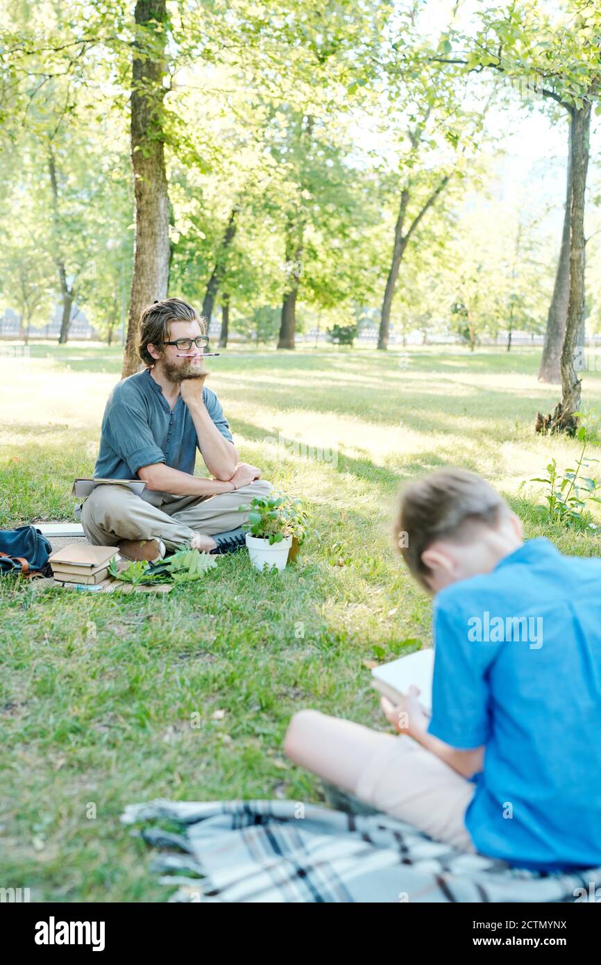 Boring young bearded teacher keeping pencil on mouth and waiting when students perform task in park Stock Photo
