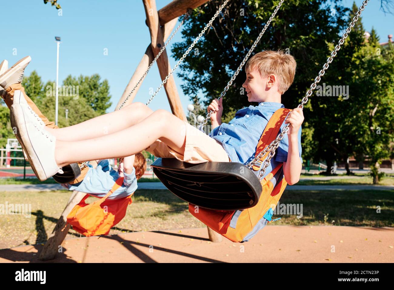 Happy school kids with satchels swinging on swings and having fun after classes Stock Photo