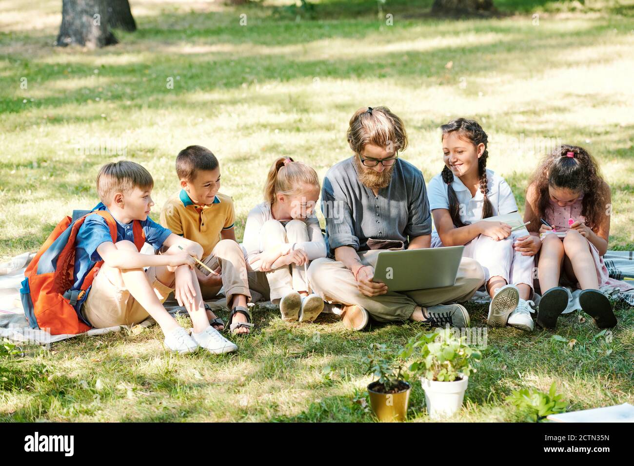 Hipster bearded teacher sitting on grass and using laptop while preparing project with schoolchildren in park Stock Photo