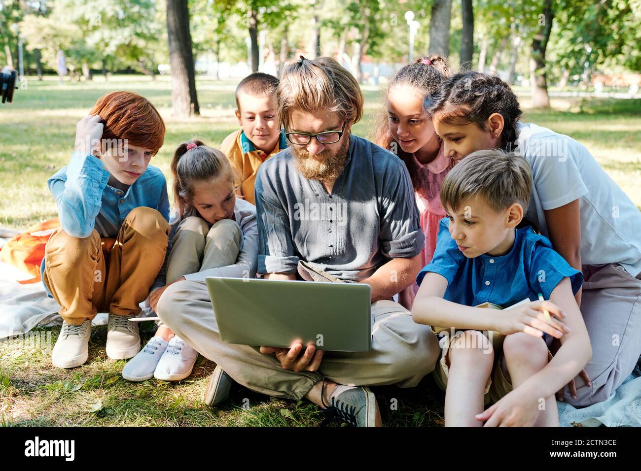 Young bearded teacher in eyeglasses sitting on grass and using laptop while showing pictures to kids at outdoor class Stock Photo