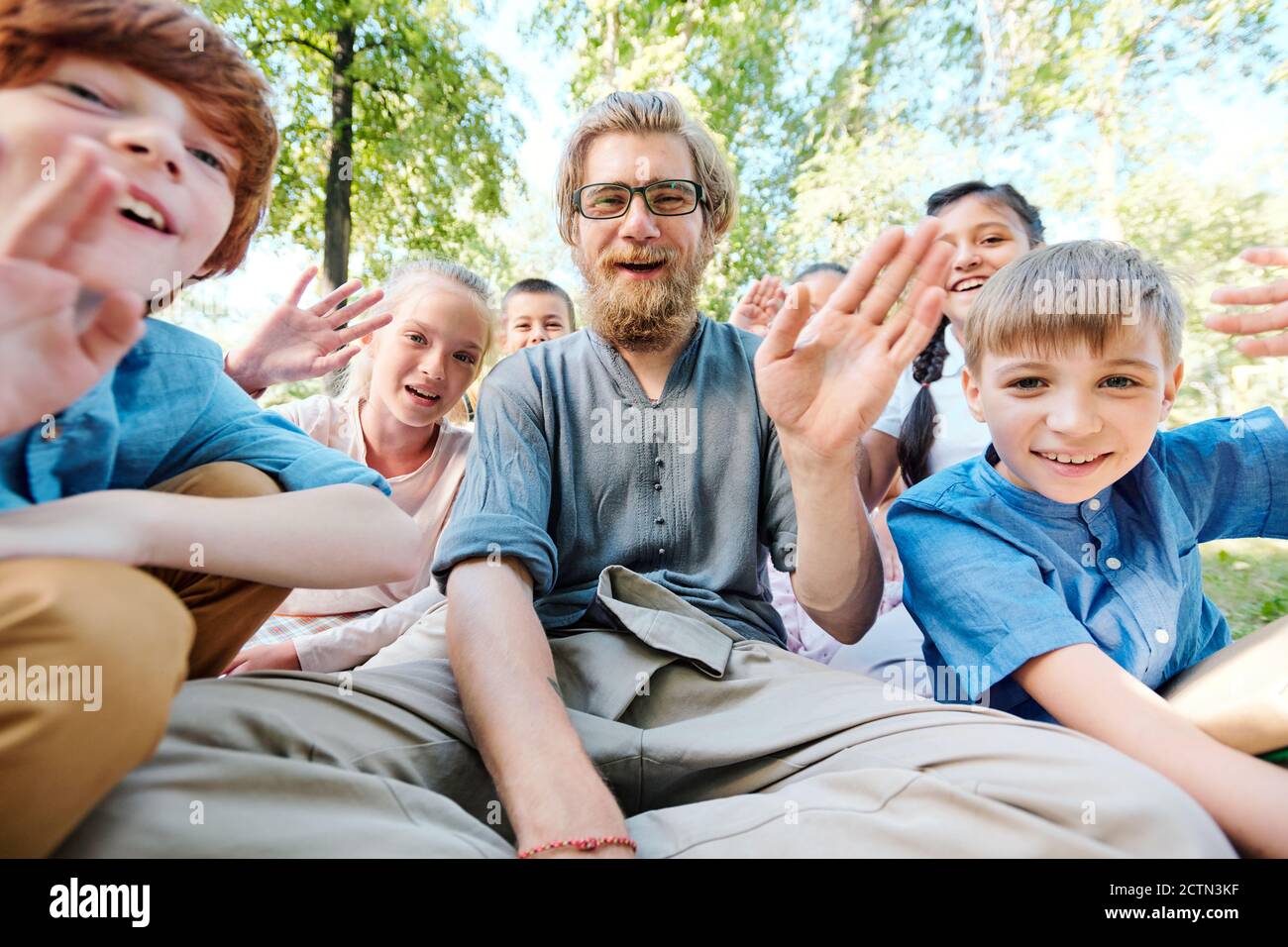 Portrait of cheerful teacher and kids sitting in park and waving hands while welcoming you during video conferencing Stock Photo