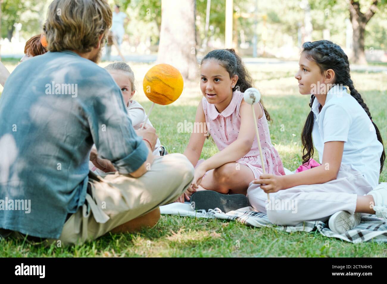 Curious multi-ethnic schoolgirls holding planet models on sticks while discussing astronomical bodies with teacher in park Stock Photo