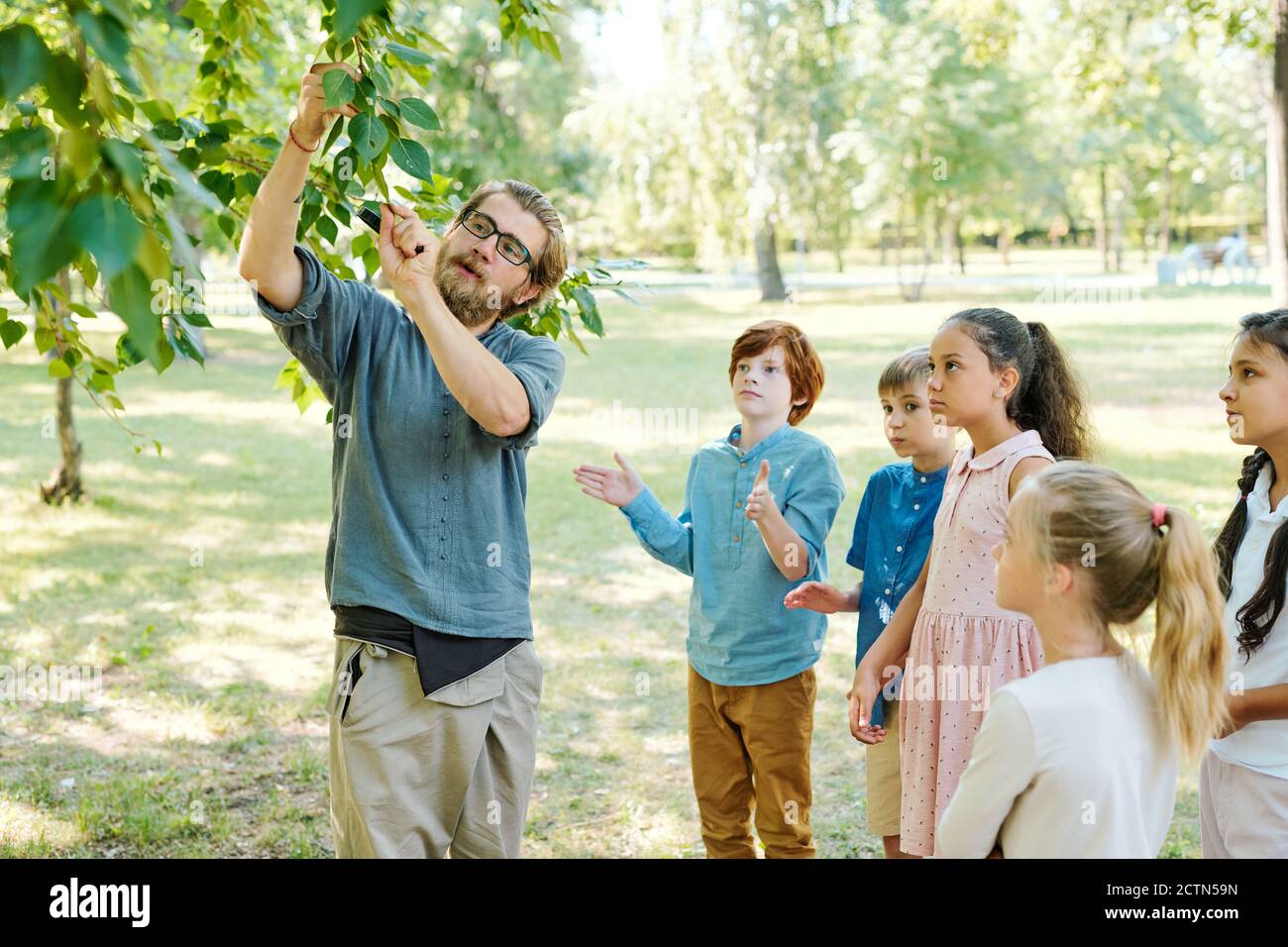 Young biology teacher in glasses standing at tree in park and showing twig to kids at outdoor class Stock Photo