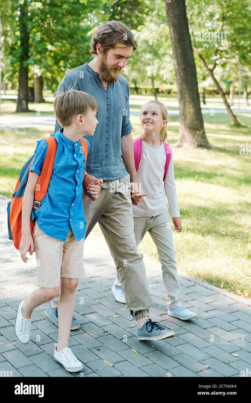 Young bearded father holding hands of kids with satchels and walking them to school across park Stock Photo