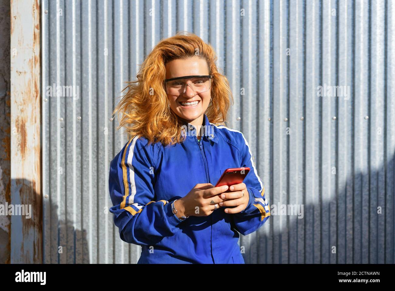 Delighted female worker in uniform and protective glasses standing near metal building and browsing cellphone while having break during work Stock Photo