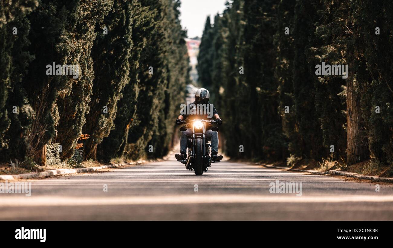 Biker in black leather jackets and helmet riding powerful motorcycle on asphalt road leading between green forest in countryside Stock Photo