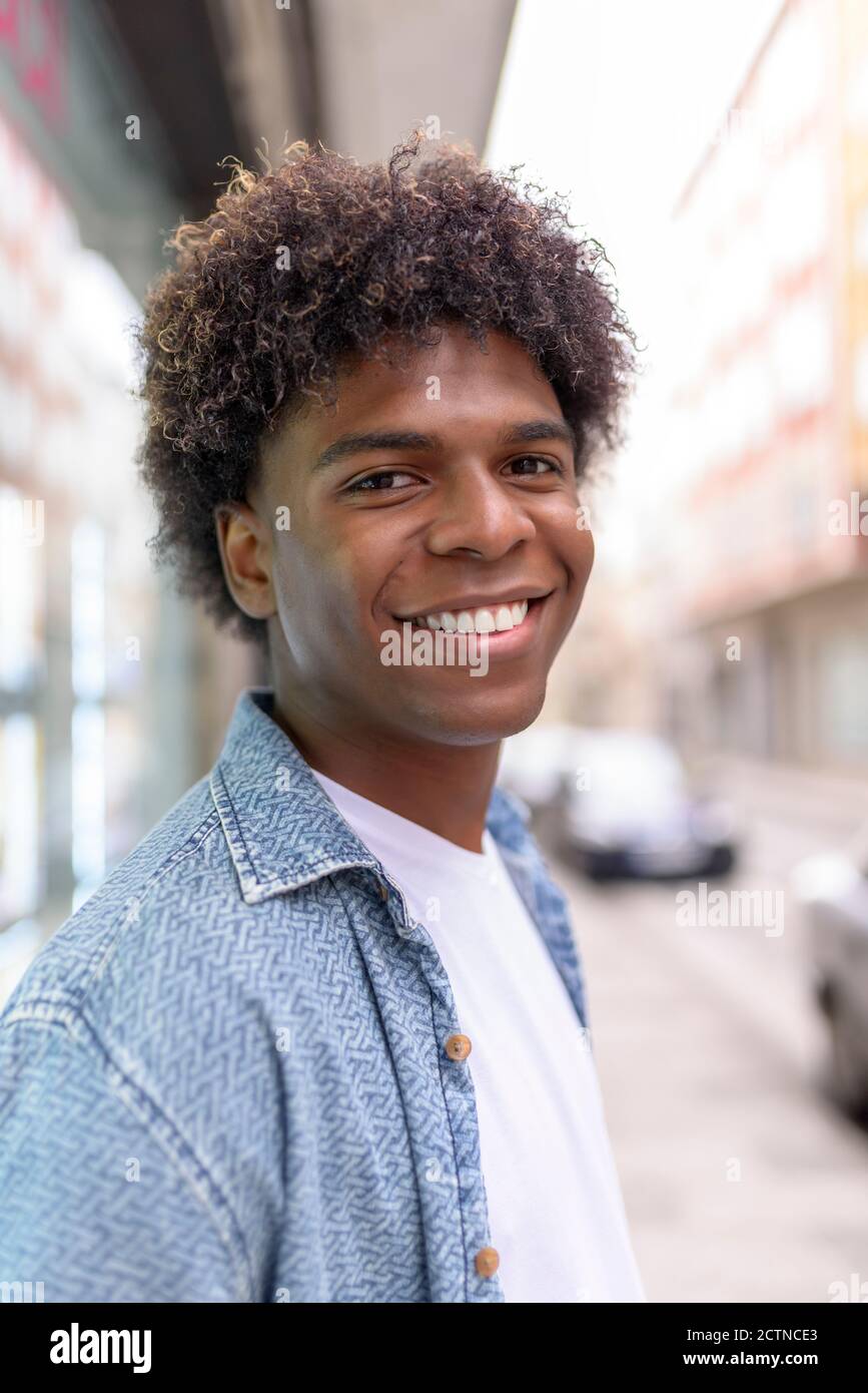 Positive African American male with Afro hairstyle and charismatic smile  looking at camera while standing on blurred background of city Stock Photo  - Alamy