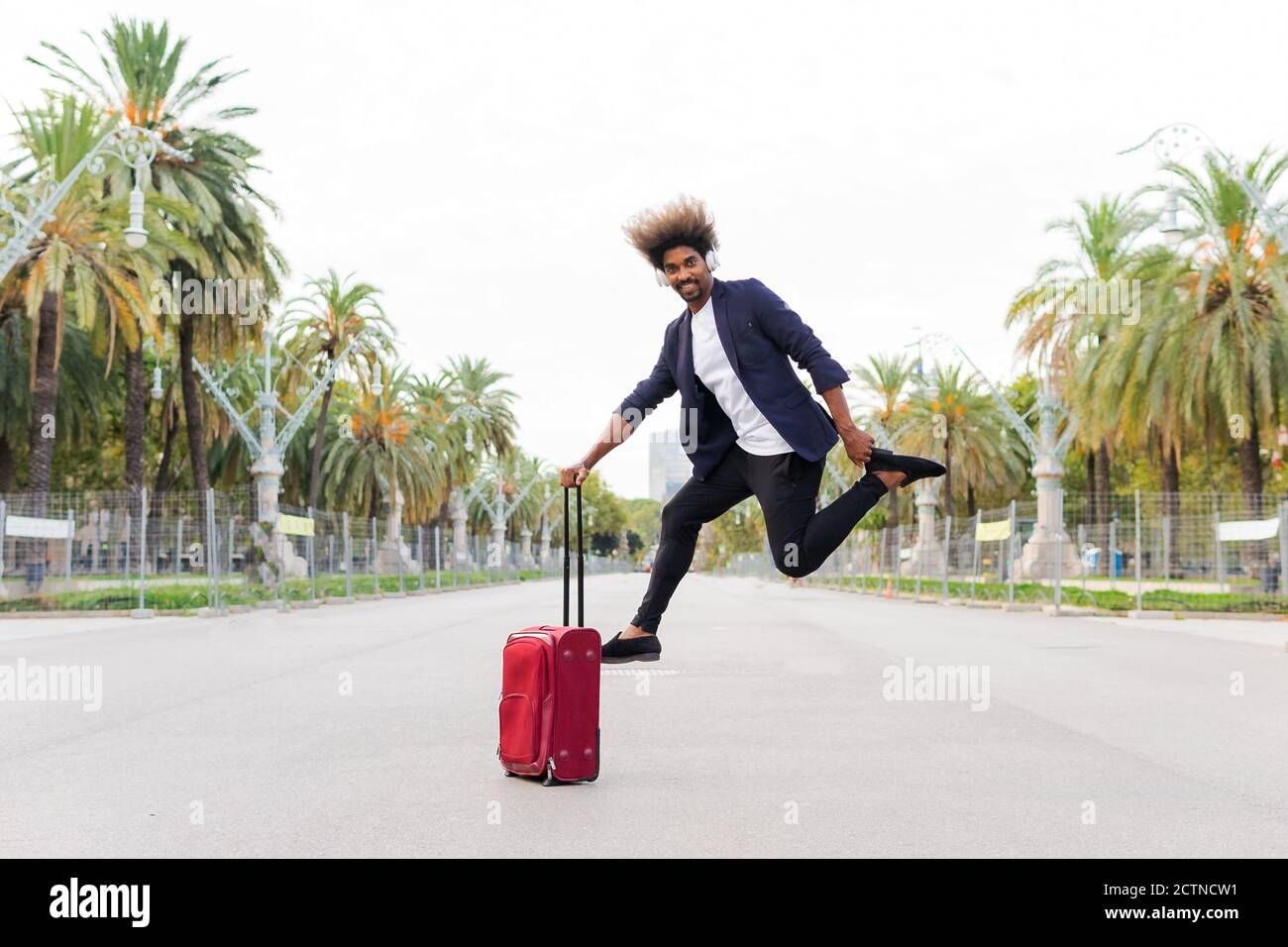 Young black man in a suit jumping and listening to music while clutching a wheeled suitcase and facing the camera on the street Stock Photo