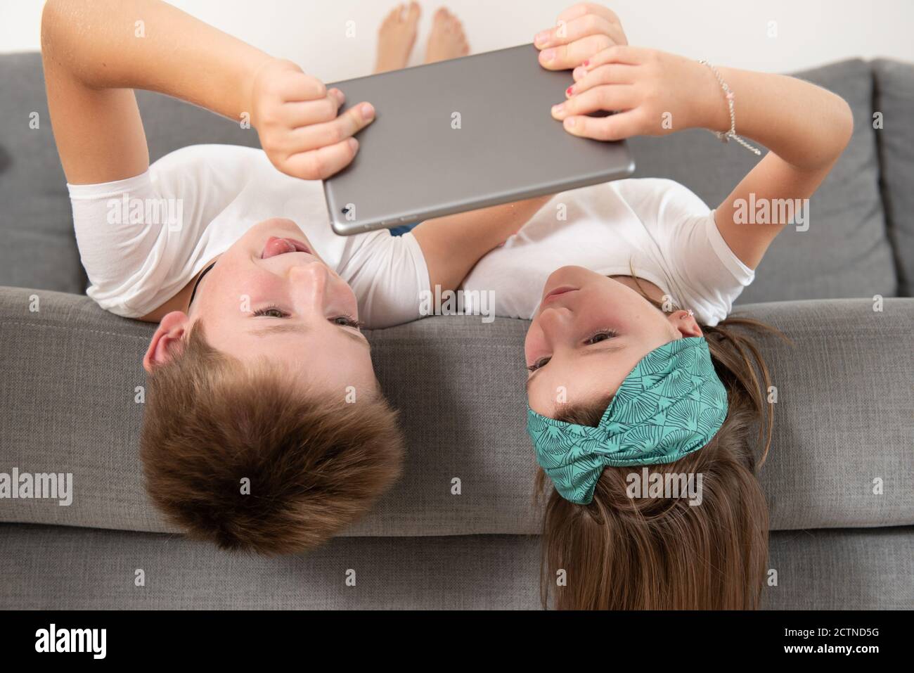Brother and sister lying face down on the couch while entertained using tablet during weekend Stock Photo