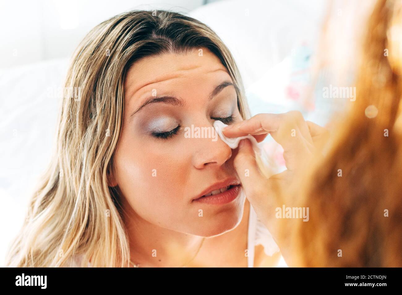 Cropped unrecognizable Woman doing makeup for female friend with eyes closed sitting on bed at home Stock Photo