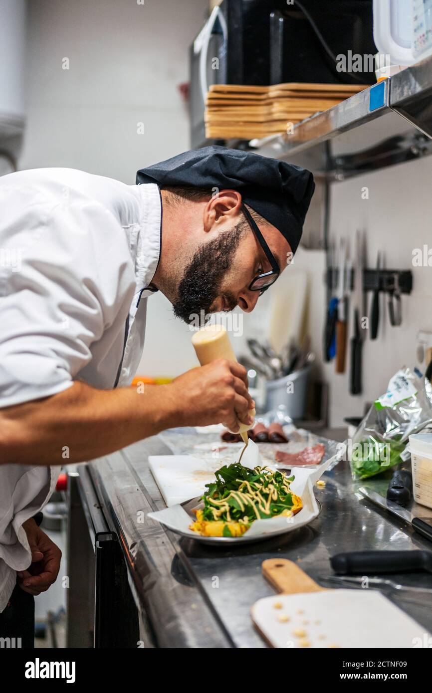 Side view of busy male chef in uniform adding sauce on top of dish with greenery while working in kitchen of cafe Stock Photo