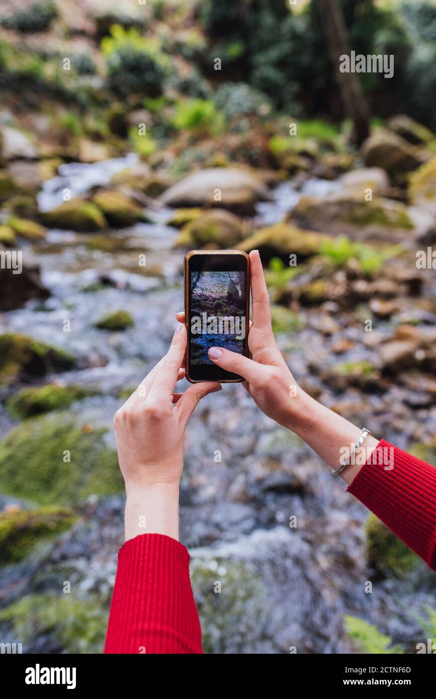Back view of anonymous female tourist sitting on shore of river in woods and taking picture of amazing landscape in Valle del Jerte Stock Photo