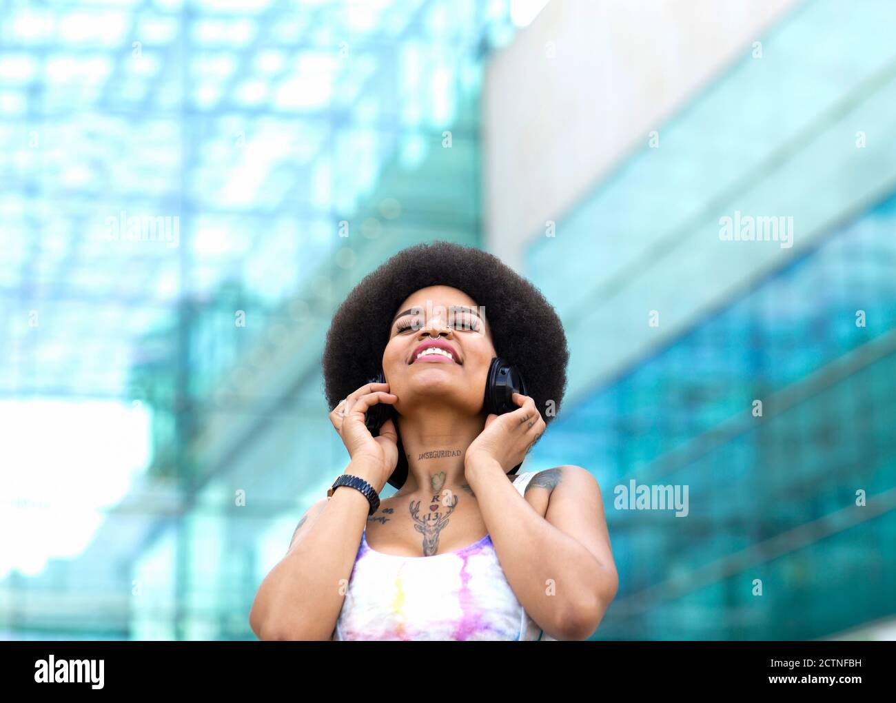 Carefree African American female listening to music in wireless headphones and enjoying songs with closed eyes while standing on street in summer Stock Photo