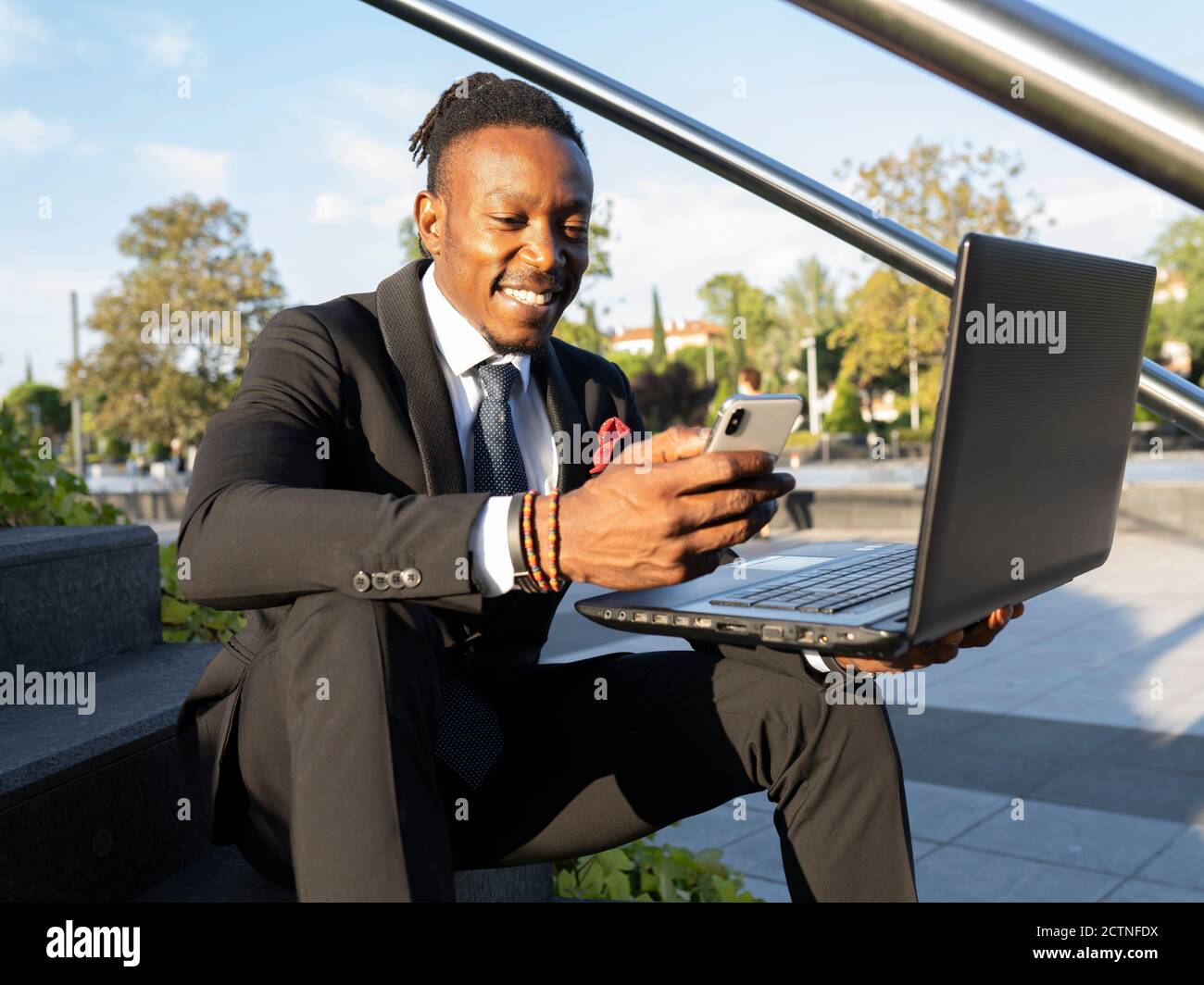 Cheerful African American businessman sitting on steps with laptop in ...