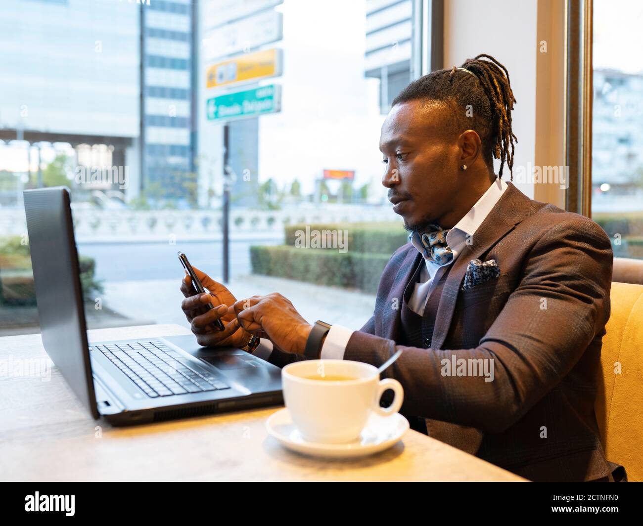 Side view of concentrated African American businessman in suit sitting at table with coffee in cafe and browsing smartphone while working on remote project Stock Photo