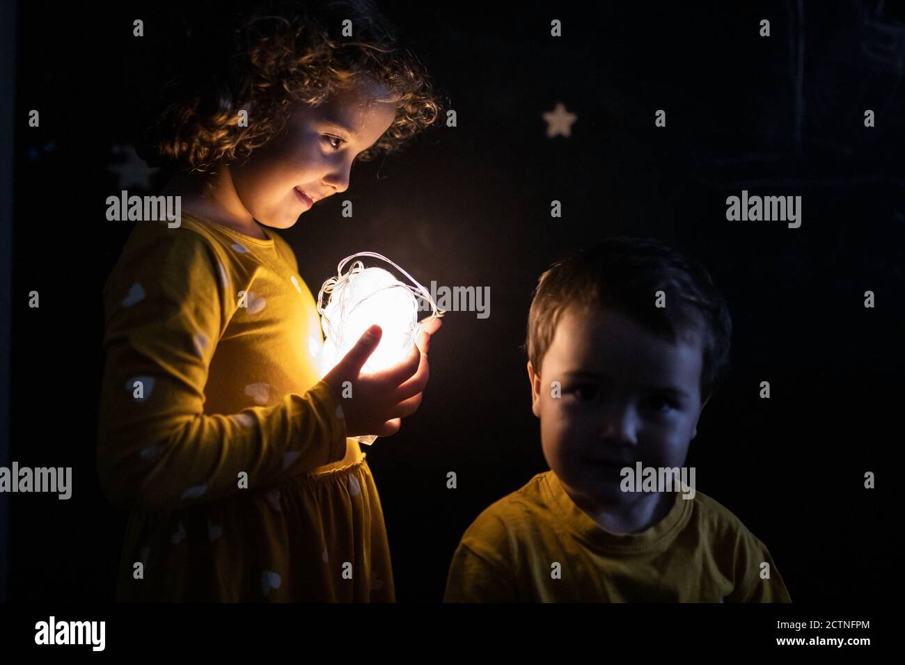 Side view of curious siblings in pajamas standing in dark room with illuminated garland Stock Photo
