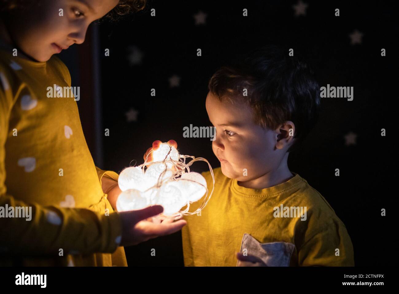 Side view of curious siblings in pajamas standing in dark room with illuminated garland Stock Photo